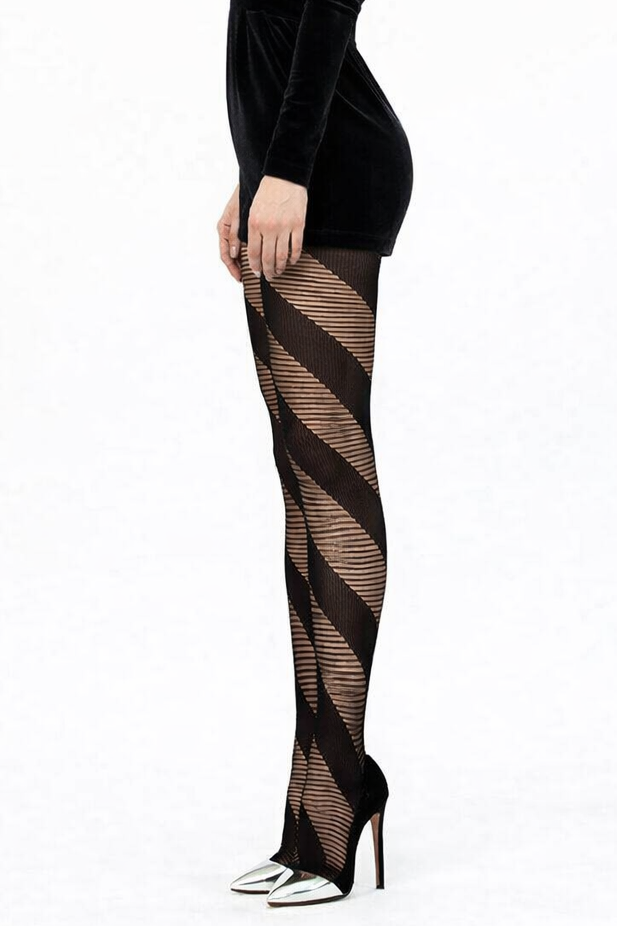 Full-length studio photo of a woman in a side pose against a white background, wearing a black velvet shorts jumpsuit with structured shoulders, sheer black diagonal-striped tights, and pointed high heels with silver cap toes. Her black hair is slicked back into a sleek ponytail, and she stands upright with a composed, fashion-model stance.