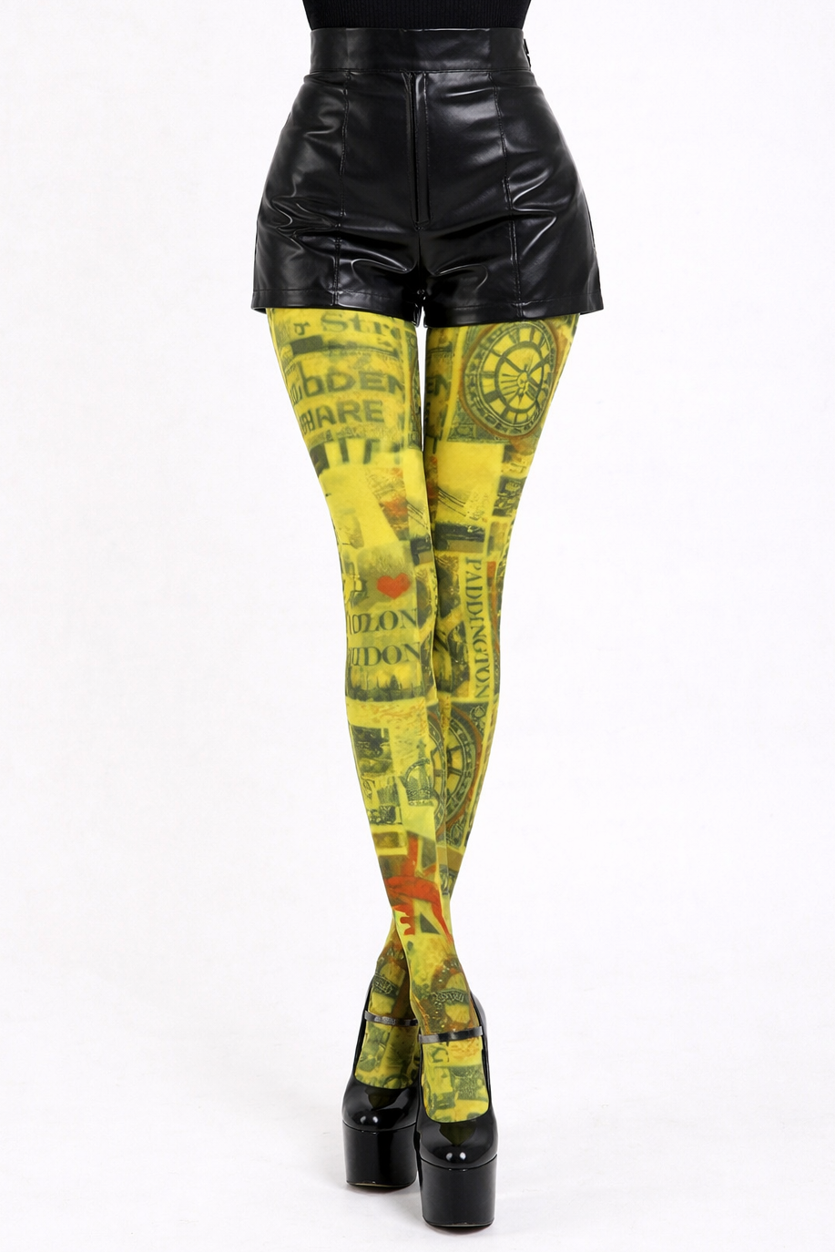 Model shown from the waist down wearing bright yellow London-print tights with legs together in a closed pose, paired with high-waisted black wet-look shorts and glossy black patent high platform heels against a plain light background.