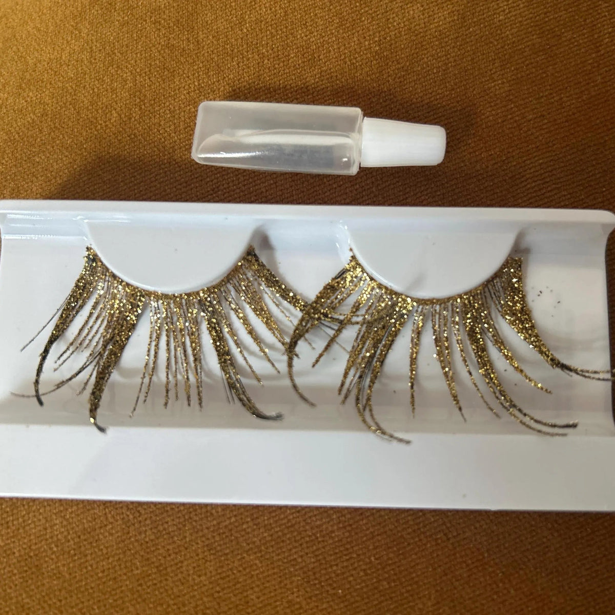 A pair of glittery gold false eyelashes in a white plastic tray with a small clear bottle above it, placed on a brown textured fabric background.