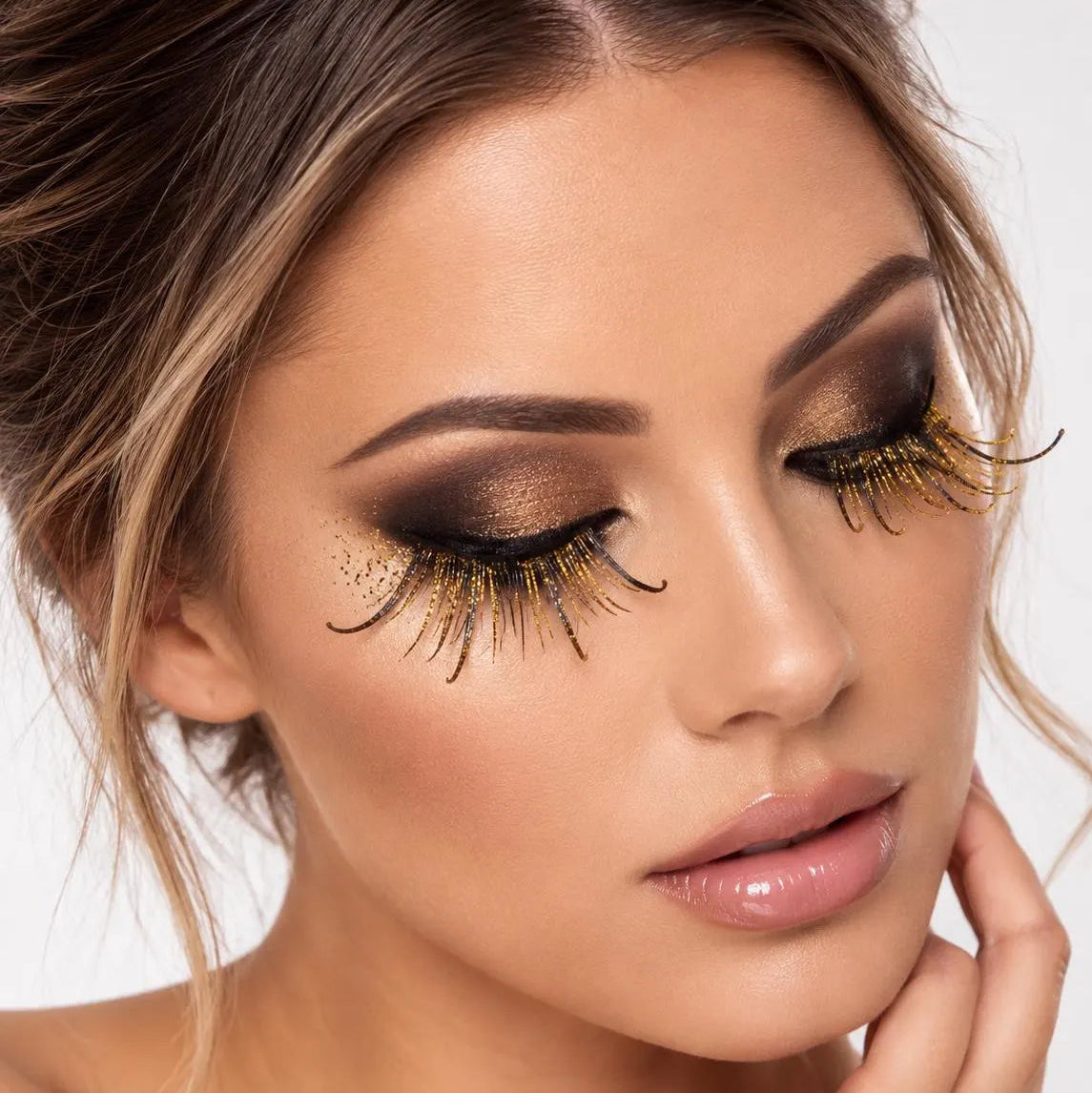 Close-up of a woman wearing dramatic false eyelashes with dark brown and gold glitter and black tips, paired with smoky eyeshadow and nude lipstick against a light gray background.