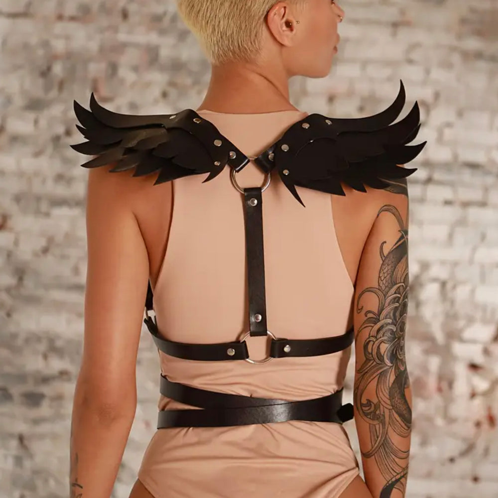 Person wearing a beige outfit with black leather harness and wing-like shoulder accessories against a stone wall.