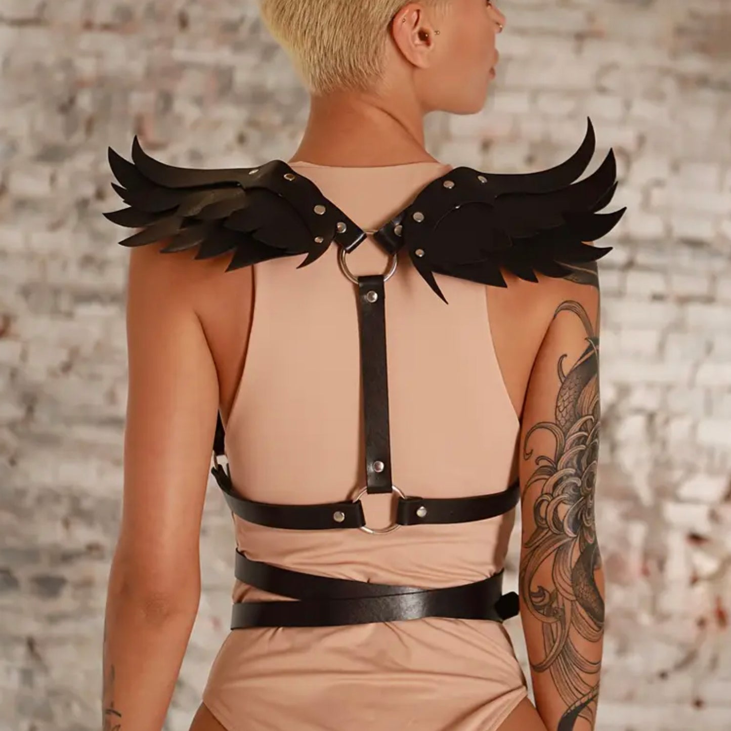 Person wearing a beige outfit with black leather harness and wing-like shoulder accessories against a stone wall.